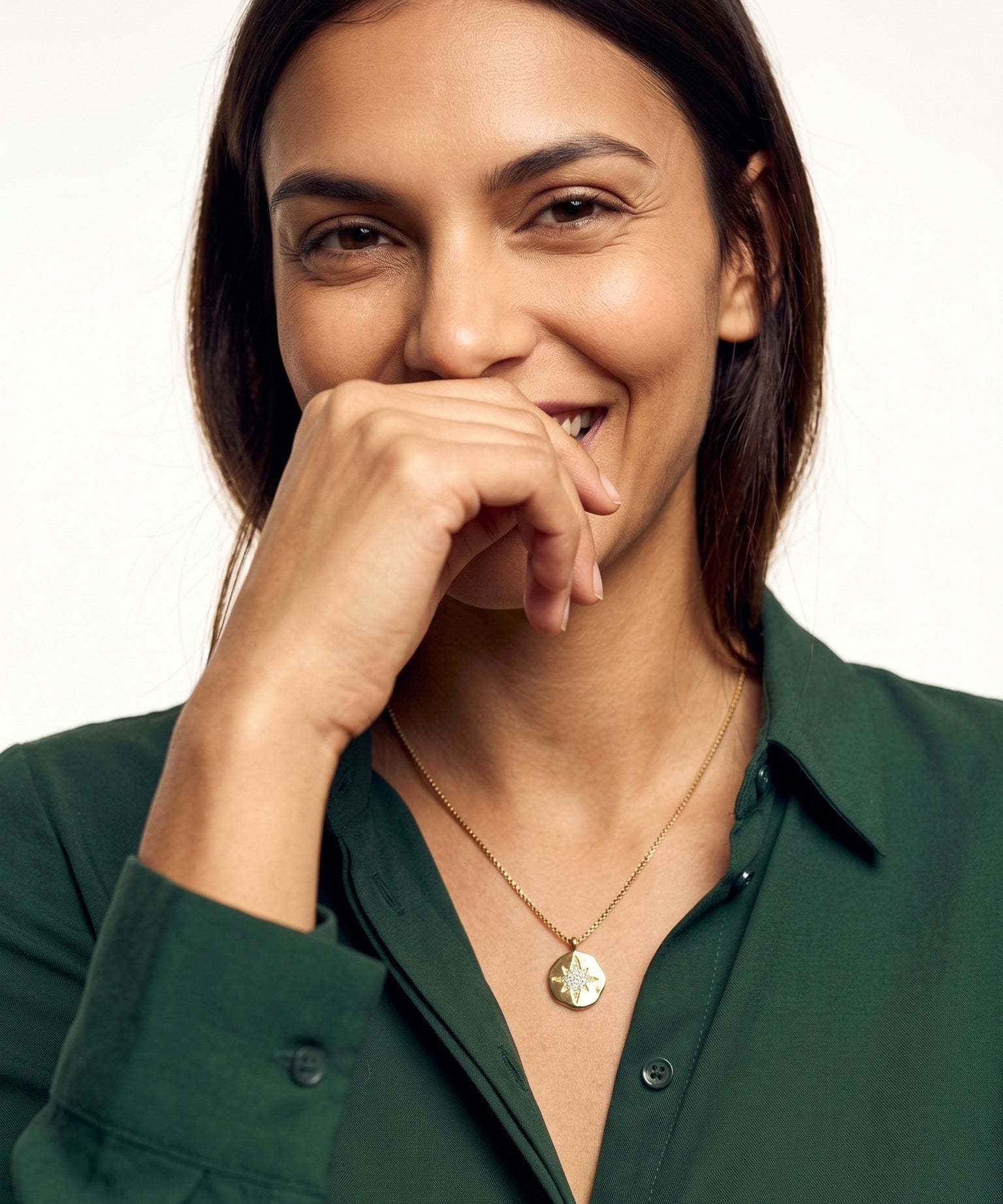 True North Necklace – worn at the chest, smiling with a hand at her mouth, gold, deep green shirt, ivory studio background