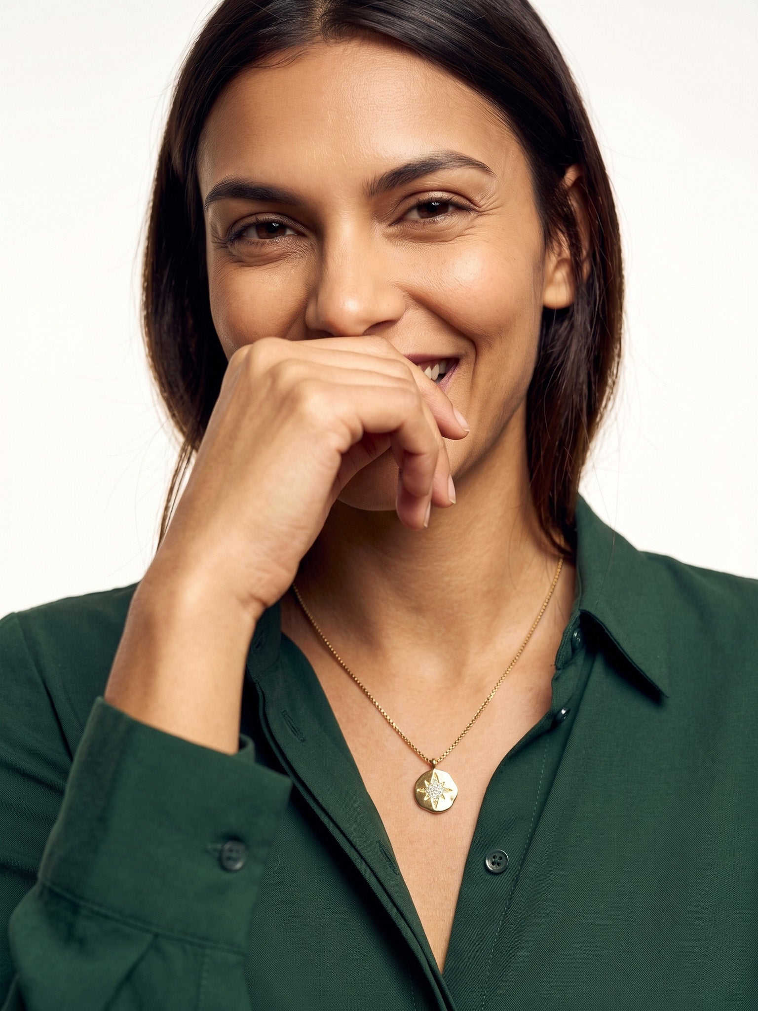 True North Necklace – worn at the chest, smiling with a hand at her mouth, gold, deep green shirt, ivory studio background