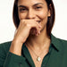 True North Necklace – worn at the chest, smiling with a hand at her mouth, gold, deep green shirt, ivory studio background