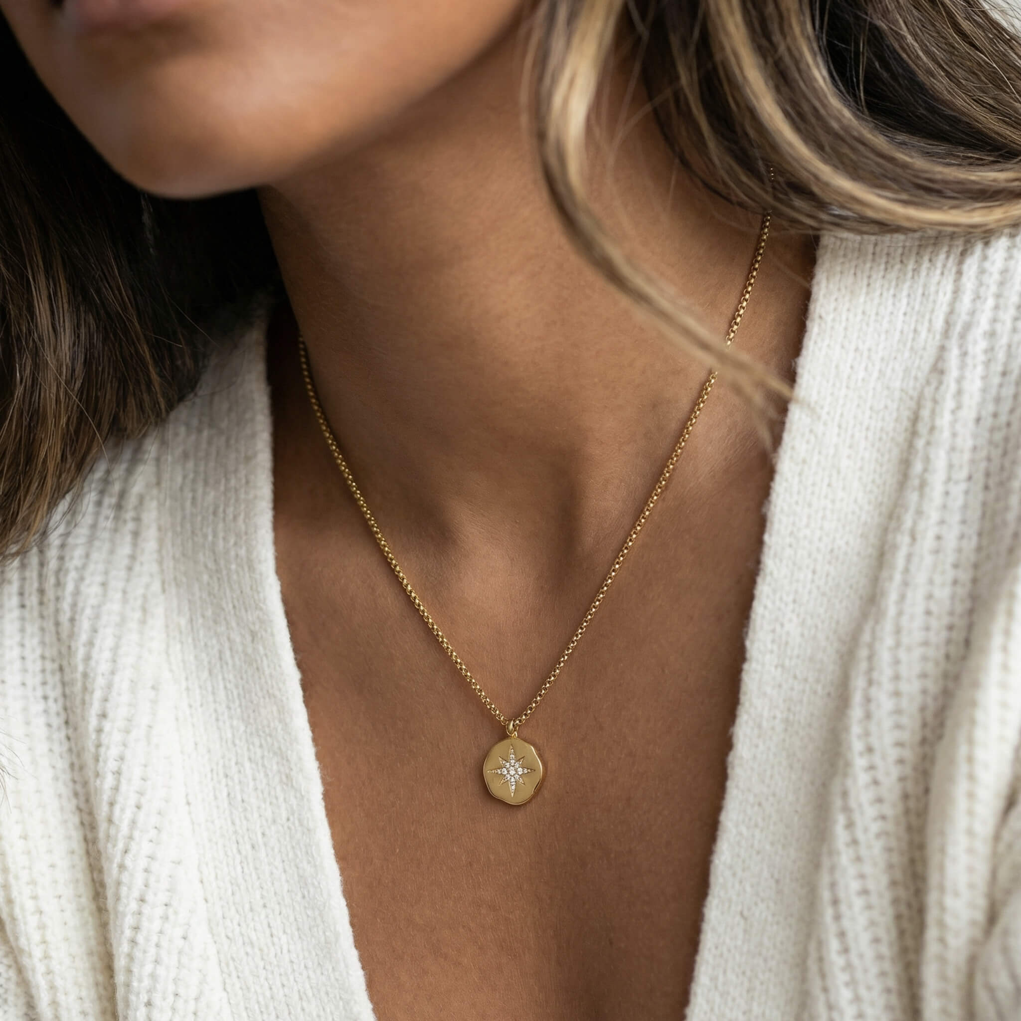 True North Necklace in gold held between two fingers, worn with a white cardigan