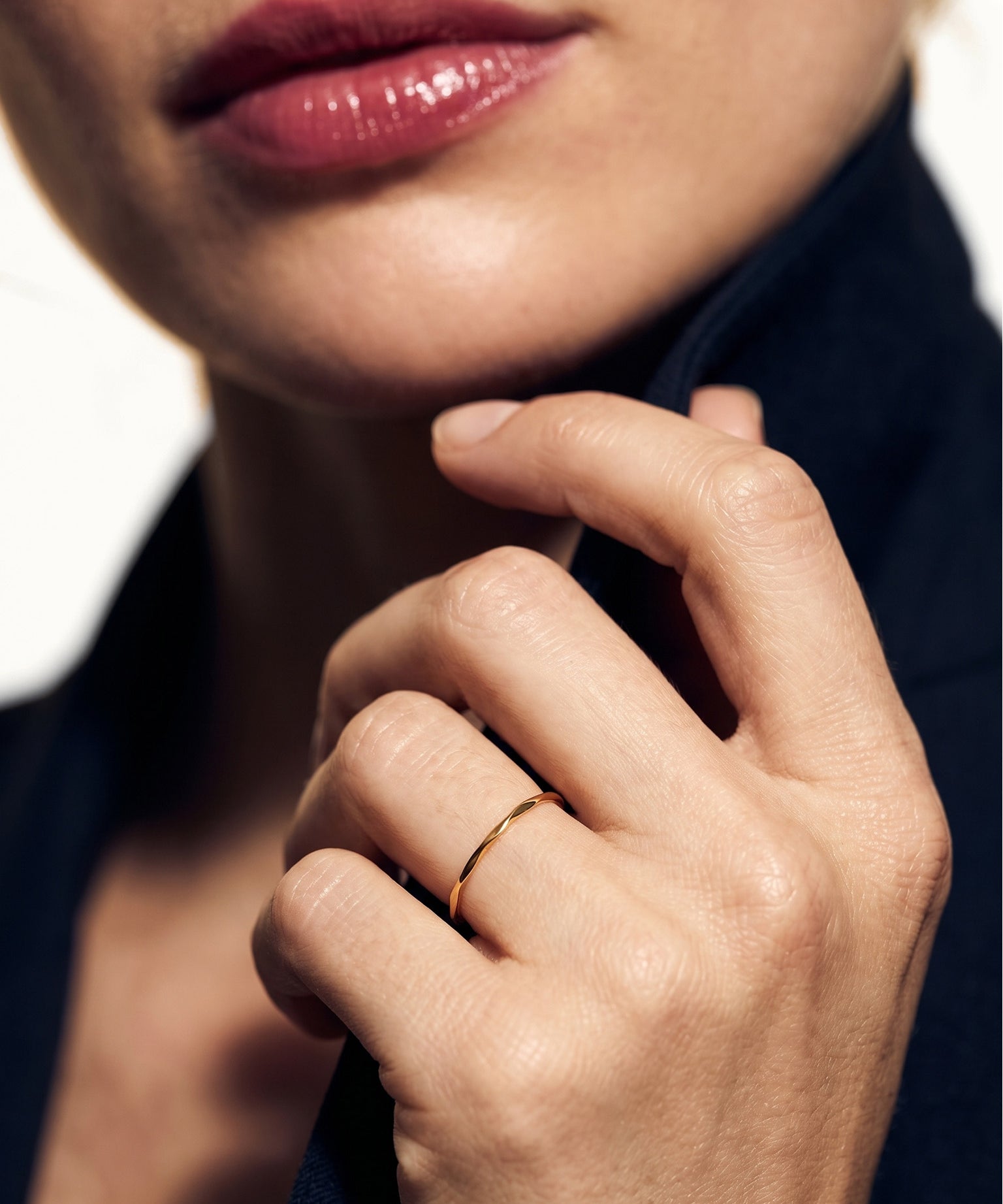 Whisper Band Ring – worn on a hand raised to the collar, close-up, gold, dark blazer, ivory studio background