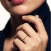 Whisper Band Ring – worn on a hand raised to the collar, close-up, gold, dark blazer, ivory studio background